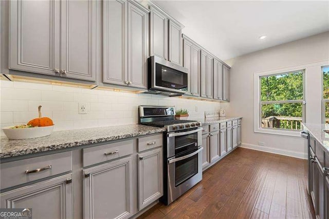 kitchen with gray cabinets, stainless steel appliances, dark hardwood / wood-style floors, light stone countertops, and decorative backsplash