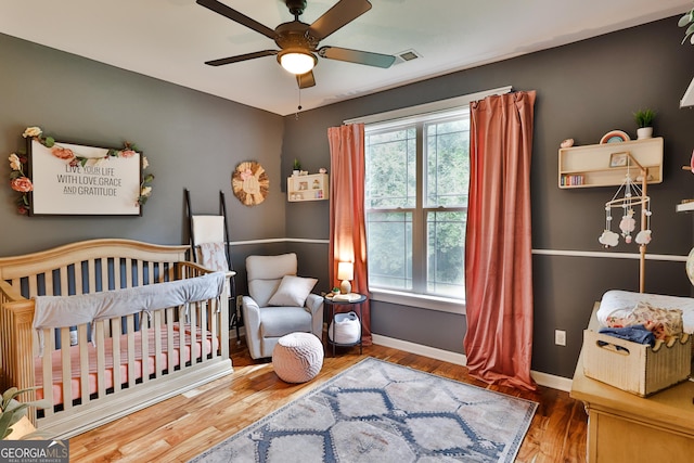 bedroom with a nursery area, ceiling fan, multiple windows, and hardwood / wood-style flooring