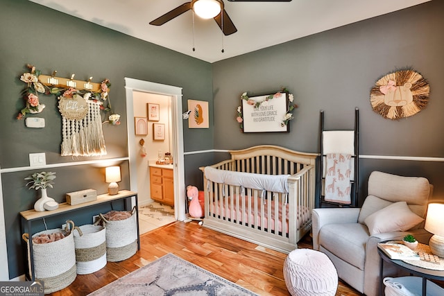 bedroom featuring wood-type flooring, a nursery area, ensuite bathroom, and ceiling fan