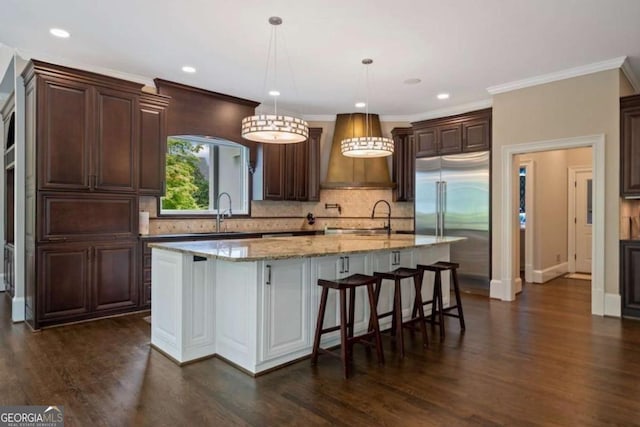 kitchen featuring sink, hanging light fixtures, stainless steel built in refrigerator, a kitchen island with sink, and custom exhaust hood