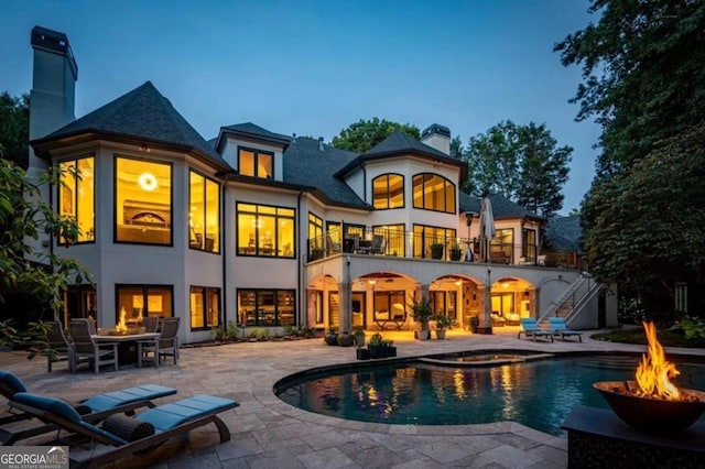 back house at dusk featuring a fire pit, a pool with hot tub, a patio area, and a balcony