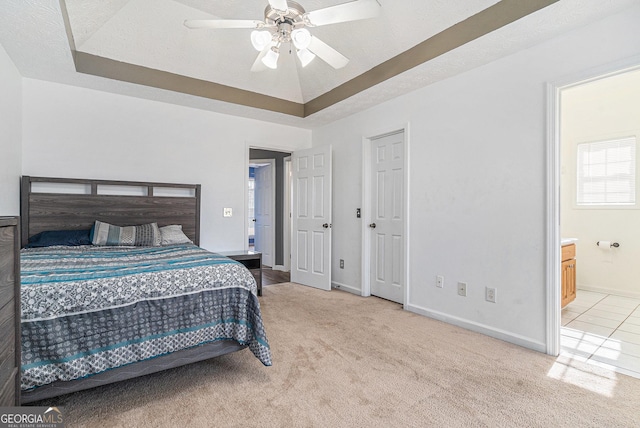 bedroom with ceiling fan, light colored carpet, ensuite bathroom, and a tray ceiling