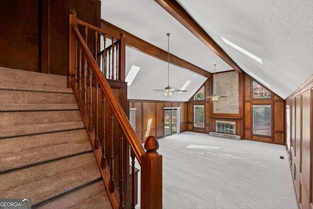 stairs featuring lofted ceiling with beams, wooden walls, a brick fireplace, carpet floors, and ceiling fan