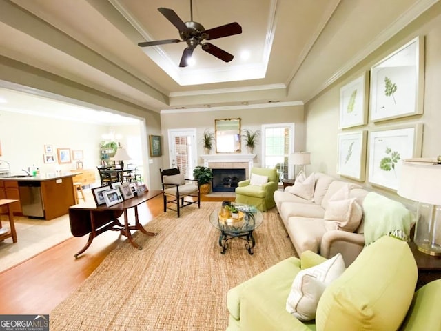 living room featuring ceiling fan with notable chandelier, sink, light wood-type flooring, a tray ceiling, and a tiled fireplace