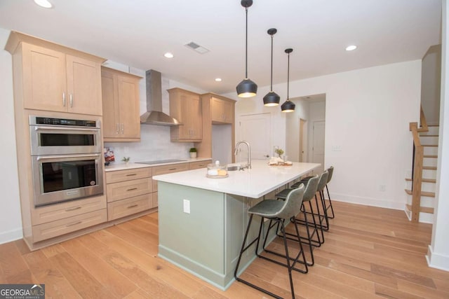 kitchen with light brown cabinets, wall chimney range hood, black electric cooktop, an island with sink, and stainless steel double oven