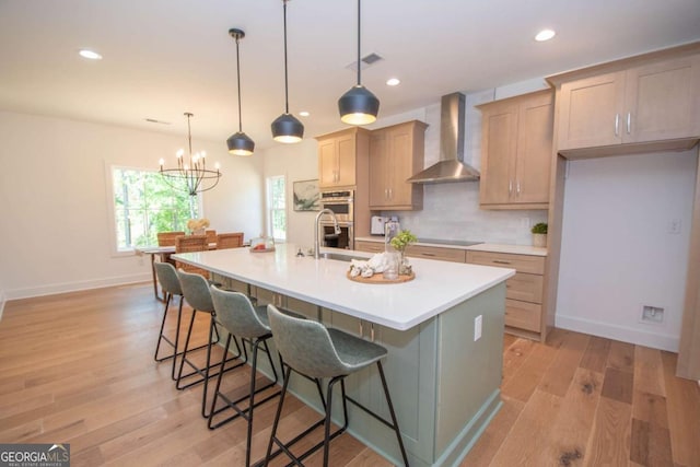 kitchen featuring light brown cabinetry, a kitchen island with sink, wall chimney range hood, a chandelier, and hanging light fixtures