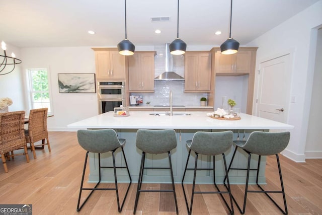 kitchen featuring a kitchen island with sink, pendant lighting, wall chimney exhaust hood, and stainless steel double oven