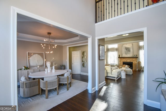 dining space with an inviting chandelier, a stone fireplace, a raised ceiling, ornamental molding, and dark hardwood / wood-style flooring