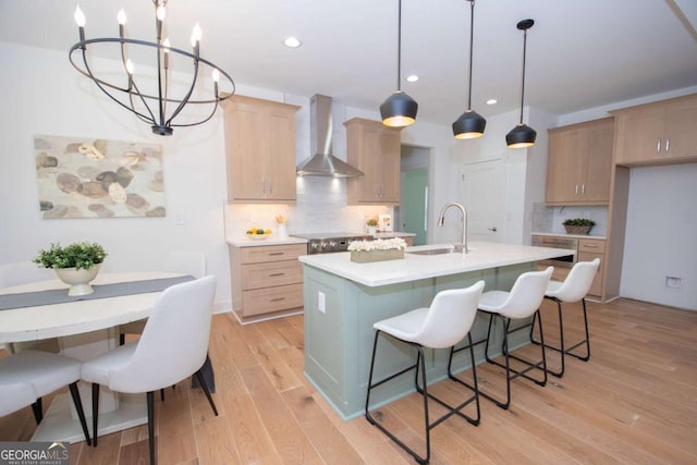 kitchen with decorative backsplash, light brown cabinetry, wall chimney range hood, pendant lighting, and an island with sink