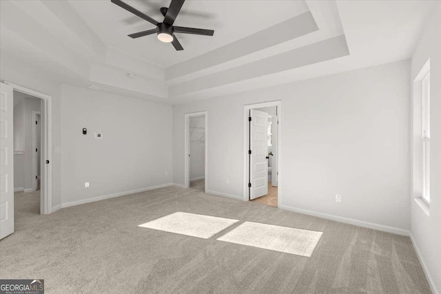 unfurnished bedroom featuring a walk in closet, ensuite bath, a tray ceiling, light colored carpet, and ceiling fan