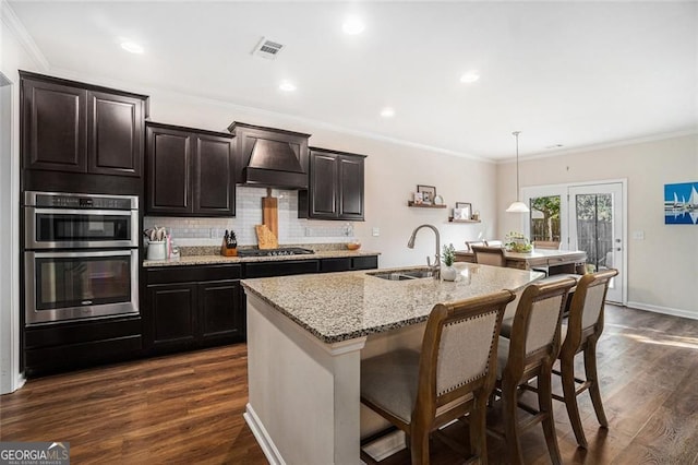 kitchen with sink, an island with sink, decorative light fixtures, custom range hood, and appliances with stainless steel finishes