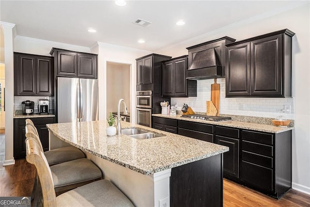 kitchen with custom range hood, light hardwood / wood-style floors, appliances with stainless steel finishes, and an island with sink