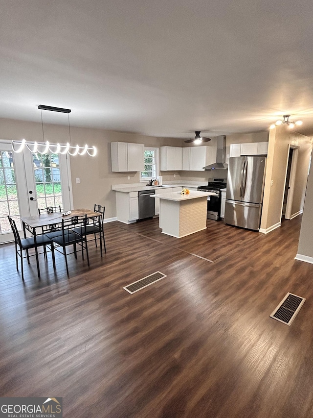 kitchen with wall chimney exhaust hood, hanging light fixtures, a kitchen island, white cabinets, and appliances with stainless steel finishes