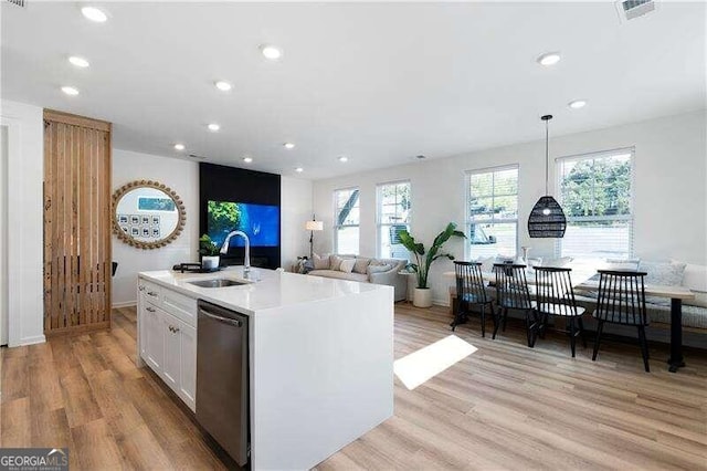 kitchen featuring stainless steel dishwasher, a kitchen island with sink, light hardwood / wood-style flooring, white cabinetry, and hanging light fixtures