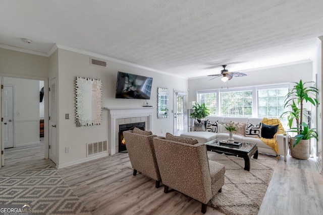 living room featuring crown molding, light hardwood / wood-style flooring, a textured ceiling, a tile fireplace, and ceiling fan