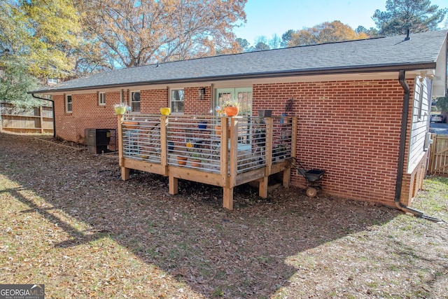 rear view of property with brick siding, roof with shingles, central AC unit, fence, and a wooden deck