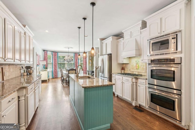 kitchen with a kitchen island with sink, sink, hanging light fixtures, light stone countertops, and stainless steel appliances