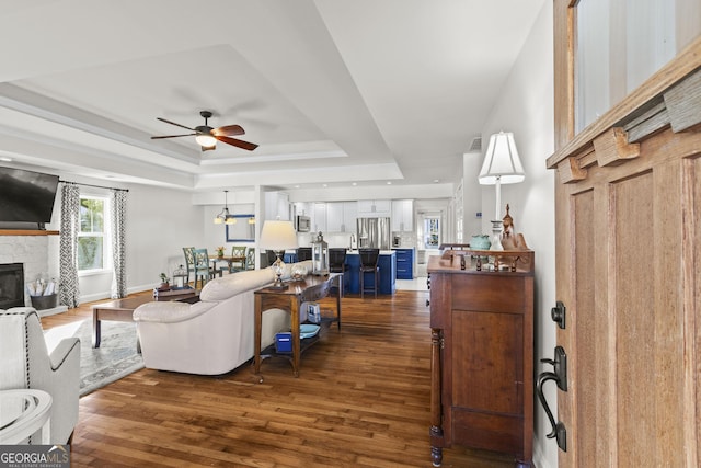 living room with a raised ceiling, a stone fireplace, ceiling fan, and dark wood-type flooring