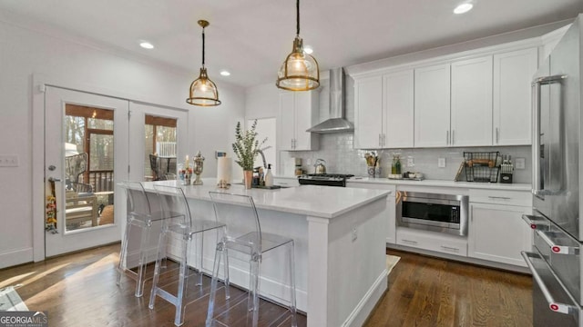 kitchen featuring pendant lighting, a kitchen island with sink, white cabinets, wall chimney exhaust hood, and appliances with stainless steel finishes