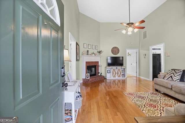 living room featuring ceiling fan, a towering ceiling, hardwood / wood-style flooring, and a brick fireplace