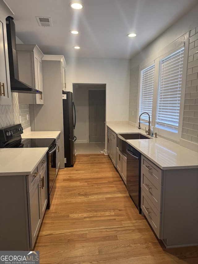 kitchen featuring gray cabinetry, wall chimney range hood, light hardwood / wood-style floors, decorative backsplash, and black appliances