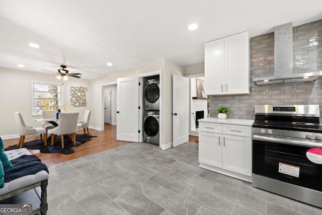 kitchen featuring white cabinetry, ceiling fan, wall chimney range hood, stainless steel range oven, and stacked washer and clothes dryer