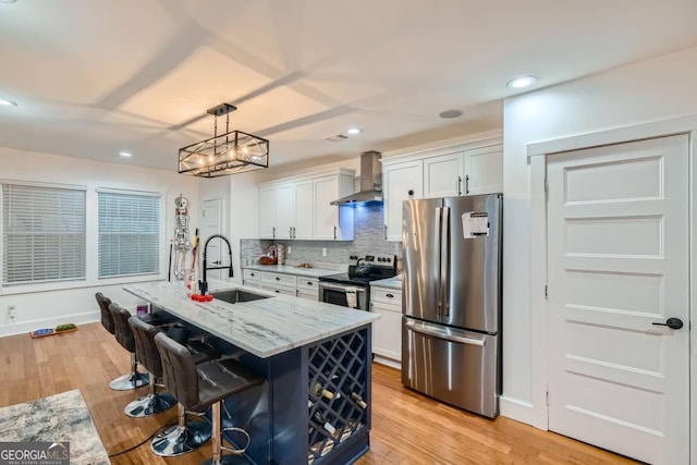 kitchen featuring white cabinetry, stainless steel appliances, wall chimney range hood, light stone counters, and a center island with sink