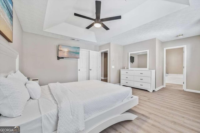 bedroom featuring light wood-type flooring, a tray ceiling, ceiling fan, and connected bathroom