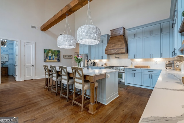 kitchen featuring beam ceiling, hanging light fixtures, an island with sink, stainless steel stove, and custom exhaust hood