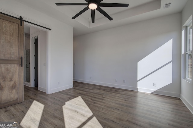 empty room featuring a raised ceiling, a barn door, ceiling fan, and dark hardwood / wood-style flooring