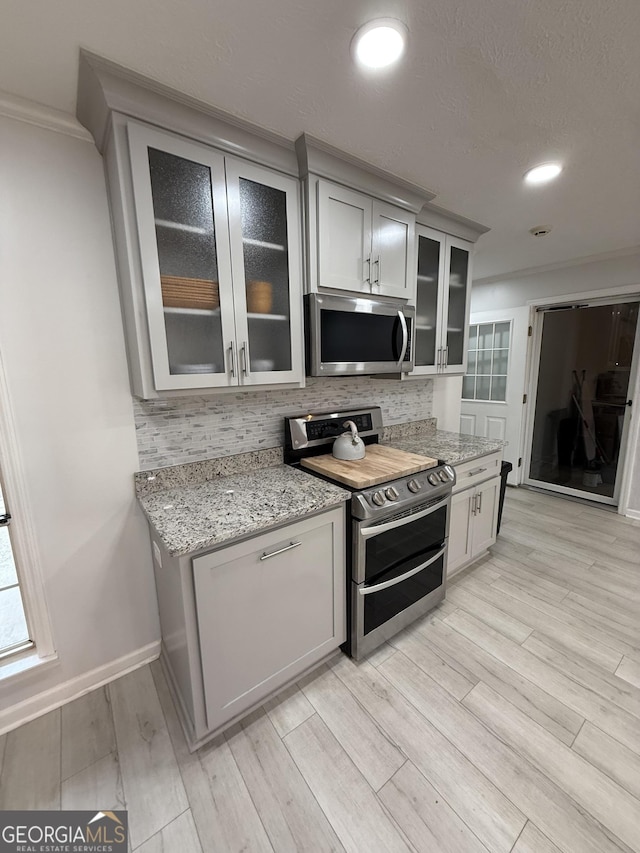 kitchen with appliances with stainless steel finishes, light wood-type flooring, backsplash, light stone counters, and ornamental molding