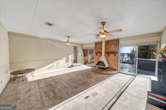 unfurnished living room featuring a wood stove, ceiling fan, carpet floors, and ornamental molding