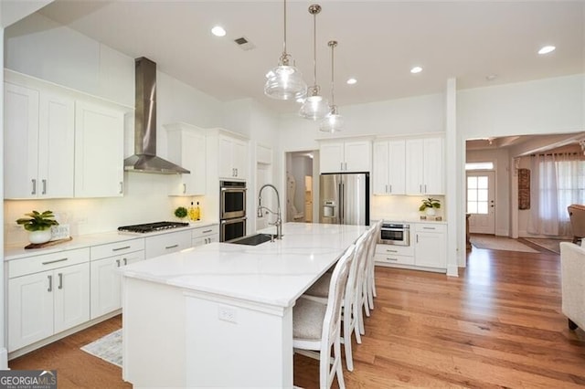 kitchen featuring a center island with sink, wall chimney range hood, appliances with stainless steel finishes, decorative light fixtures, and white cabinetry