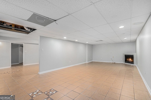 finished below grade area featuring a paneled ceiling, light tile patterned floors, a warm lit fireplace, and baseboards