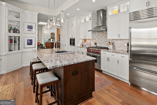 kitchen with white cabinets, wall chimney range hood, sink, an island with sink, and premium appliances