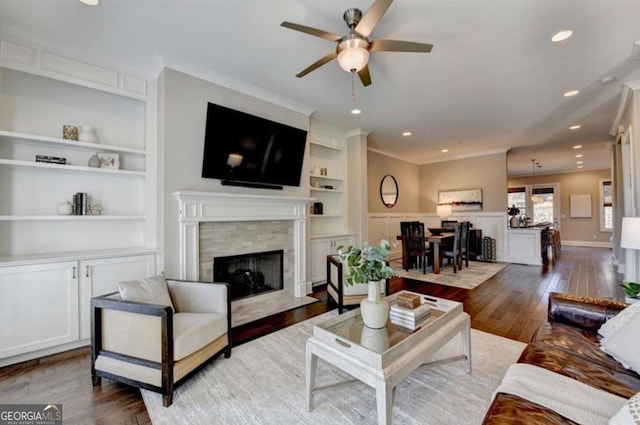 living room with built in shelves, wood-type flooring, ceiling fan, and crown molding