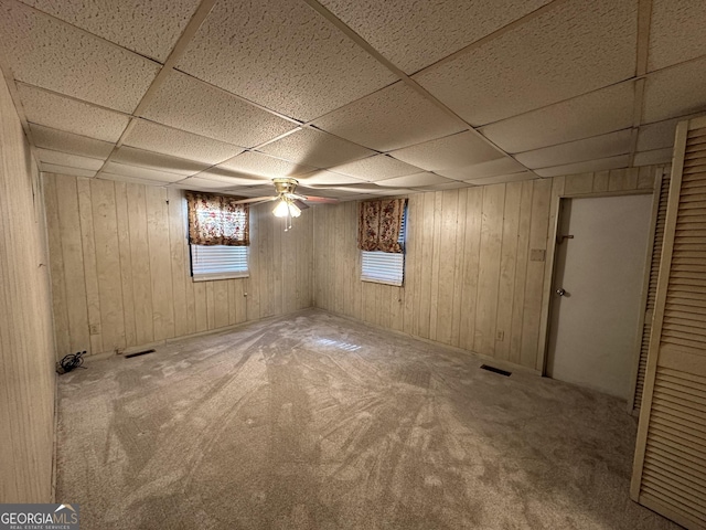 basement featuring carpet flooring, a drop ceiling, wooden walls, and ceiling fan