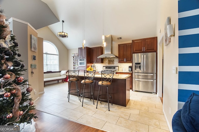 kitchen with a breakfast bar, stainless steel appliances, wall chimney range hood, a center island with sink, and hanging light fixtures