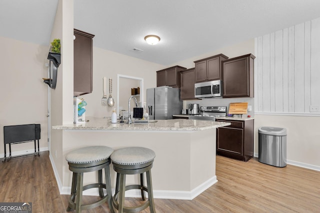 kitchen with dark brown cabinetry, light stone countertops, light hardwood / wood-style floors, kitchen peninsula, and stainless steel appliances