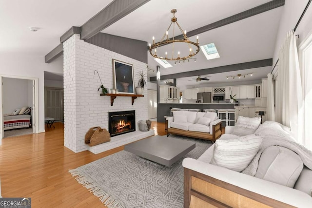 living room featuring ceiling fan with notable chandelier, light hardwood / wood-style floors, a brick fireplace, and vaulted ceiling with skylight