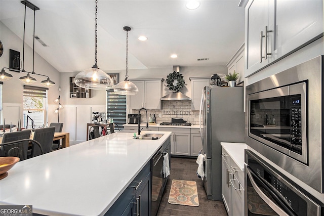 kitchen featuring sink, wall chimney exhaust hood, stainless steel appliances, pendant lighting, and a kitchen island with sink