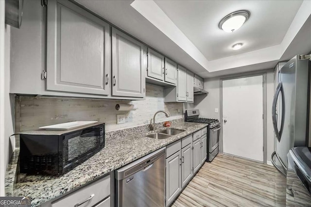 kitchen featuring light stone countertops, appliances with stainless steel finishes, gray cabinetry, a tray ceiling, and sink