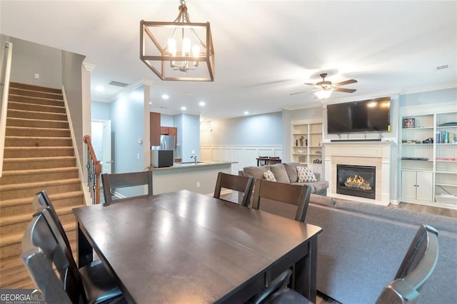 dining room with ceiling fan with notable chandelier, built in features, wood-type flooring, and ornamental molding