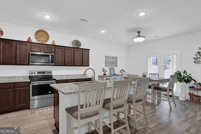 kitchen with stainless steel appliances, sink, an island with sink, light hardwood / wood-style flooring, and light stone countertops