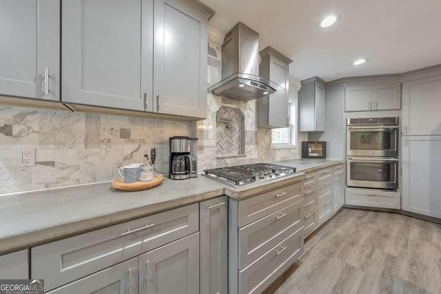 kitchen featuring appliances with stainless steel finishes, wall chimney exhaust hood, decorative backsplash, gray cabinets, and light hardwood / wood-style flooring