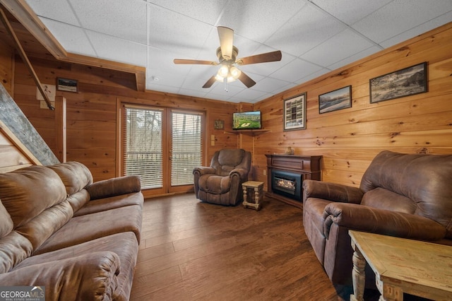 living room with dark hardwood / wood-style flooring, a drop ceiling, wood walls, and ceiling fan