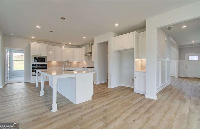 kitchen featuring white cabinets, oven, wall chimney range hood, built in microwave, and an island with sink