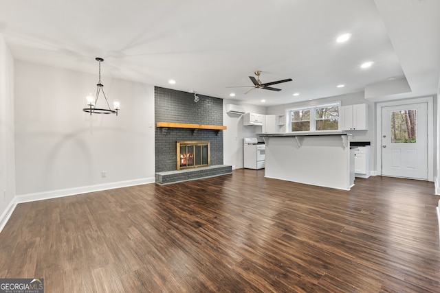 unfurnished living room with ceiling fan with notable chandelier, an AC wall unit, a brick fireplace, and dark wood-type flooring