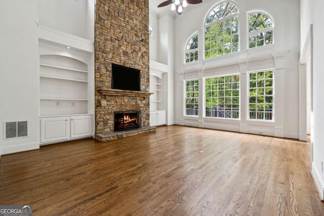 unfurnished living room with ceiling fan, a healthy amount of sunlight, a stone fireplace, built in features, and a towering ceiling