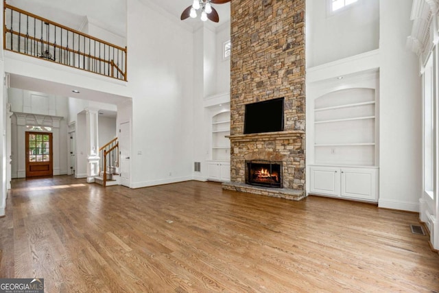 unfurnished living room featuring light wood-type flooring, ceiling fan, built in features, a high ceiling, and a stone fireplace